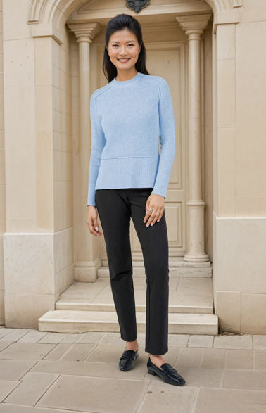 Woman wearing a light blue sweater and black pants standing in front of a stone building.