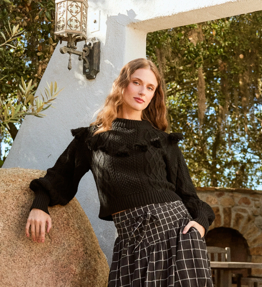 woman wearing a black knit sweater, textured with tassel details around the top, lifestyle shot, paired with a checkered skirt, and leaning on a rock