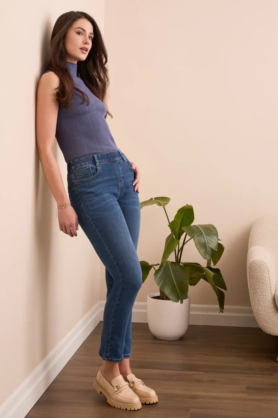 Woman wearing a blue top and jeans leaning against a wall with a plant in the background