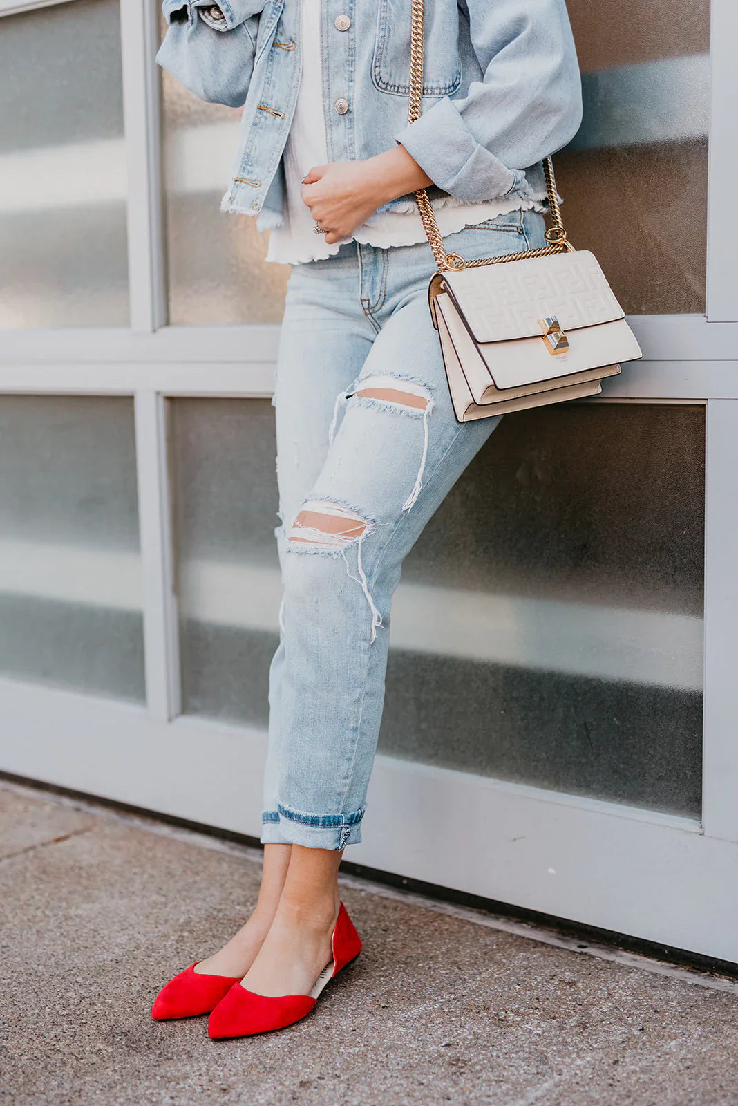 Person wearing a light blue denim jacket, ripped jeans, and red flats, holding a beige handbag against a glass wall.