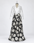 Connie Roberson Greyson Amazon Wrap Skirt A long formal dress displayed on a mannequin against a light gray studio background. The dress has a white satin long-sleeve button-front bodice and a full black skirt with large silver floral embroidery.
