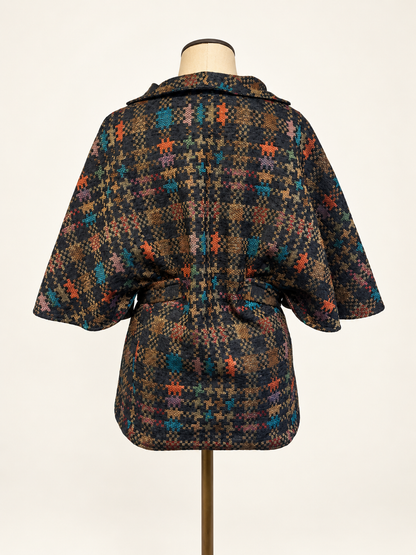A short hooded cape-style jacket with a tie waist belt, displayed on a dress form. The fabric features a dense multicolor plaid and houndstooth pattern with black, brown, orange, and teal tones.