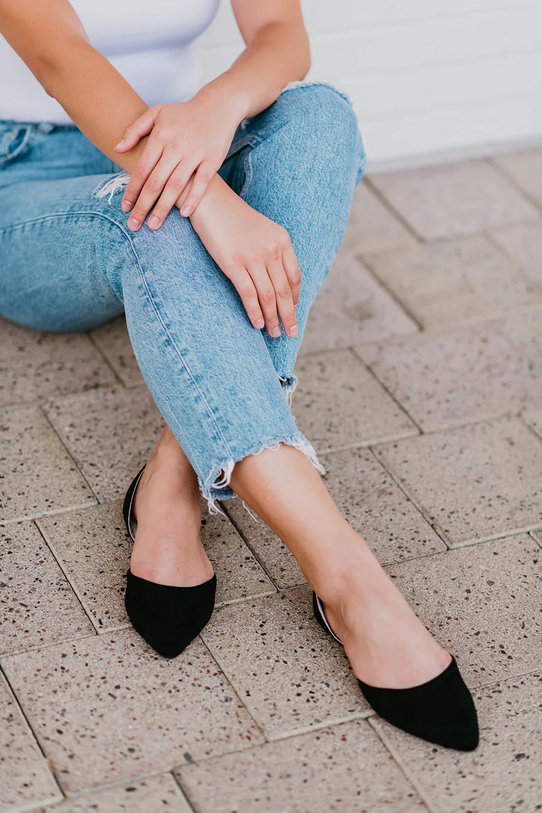 Person wearing black pointed-toe shoes and light blue jeans sitting on a stone pavement.
