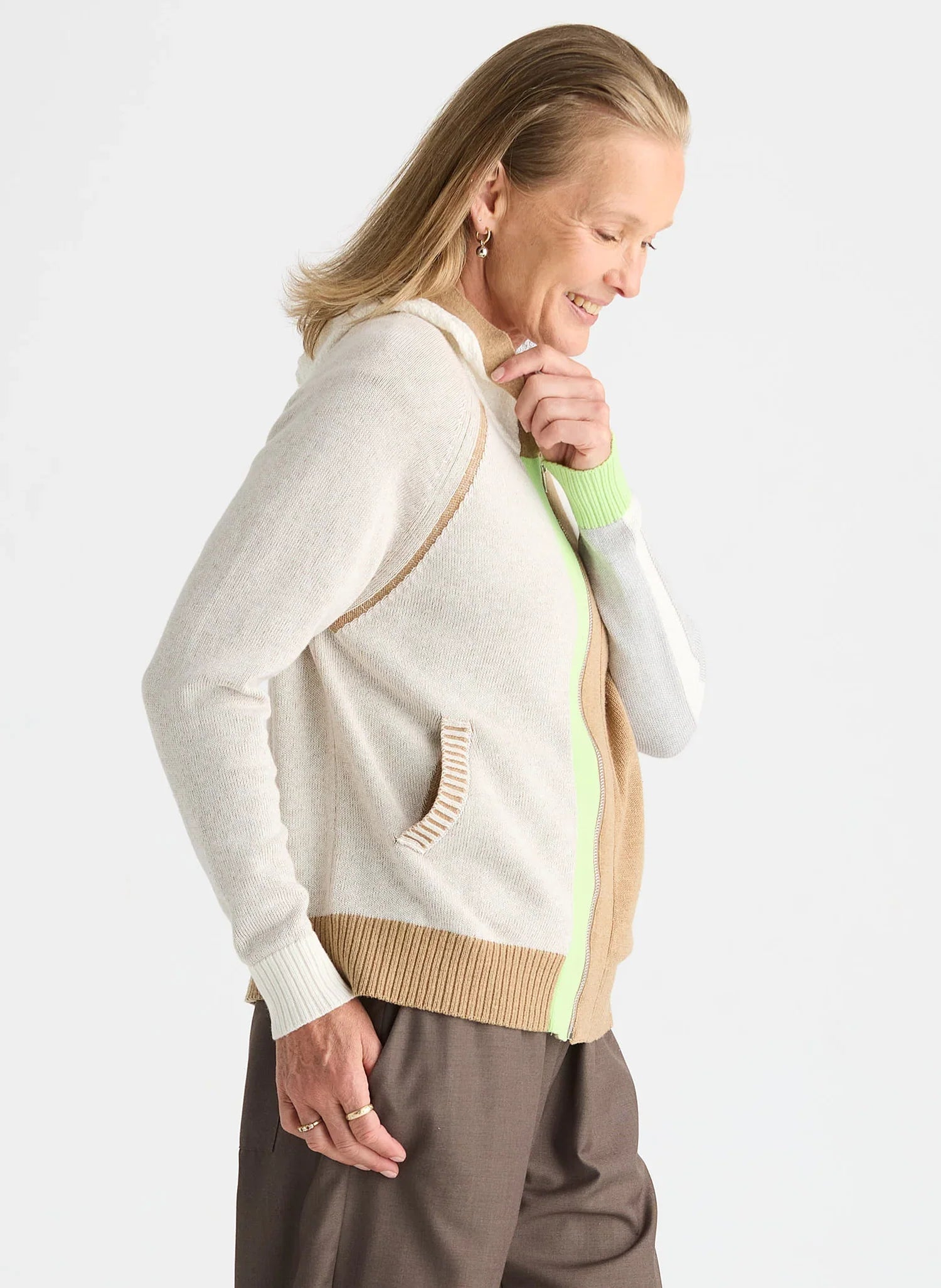 A woman wearing a cream-colored knit cardigan with beige and lime green color-blocked details, paired with brown pants, photographed against a light gray studio background.