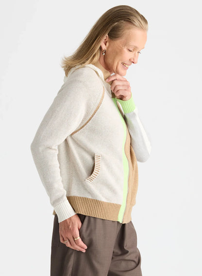 A woman wearing a cream-colored knit cardigan with beige and lime green color-blocked details, paired with brown pants, photographed against a light gray studio background.