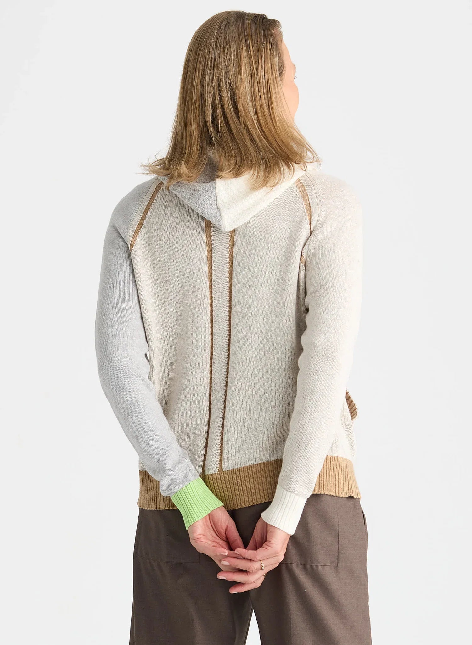A woman shown from the back wearing a light beige and gray knit hooded sweater with brown pants, against a plain light gray studio background.