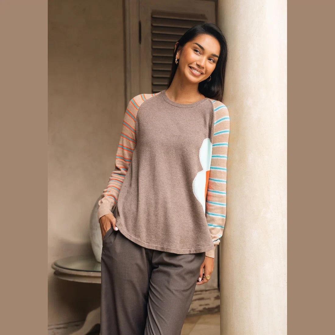 A woman modeling a long-sleeve crewneck top with a solid brown body, peach and turquoise striped sleeves, and a large light blue and coral abstract flower on the right side Zaket &amp; Plover Walnut Bright Side Sweater