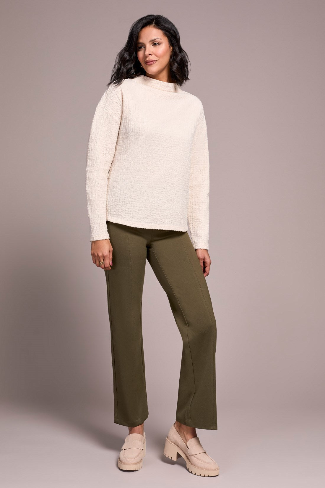 Woman wearing a cream sweater and green pants against a plain background
