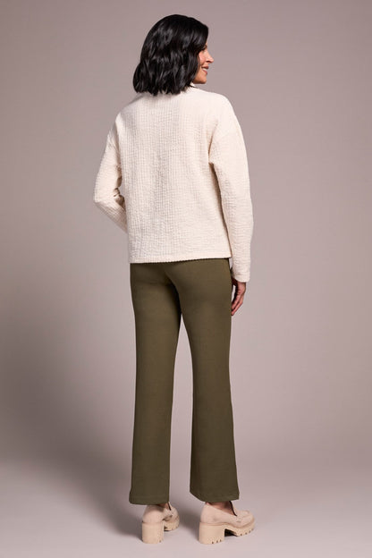 Woman wearing a cream sweater and green pants against a plain background