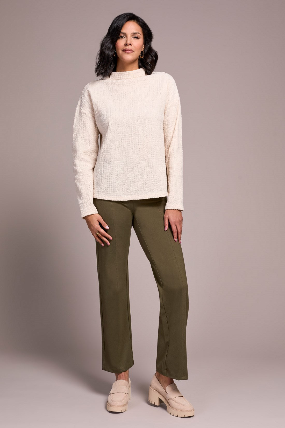 Woman wearing a cream sweater and green pants against a plain background