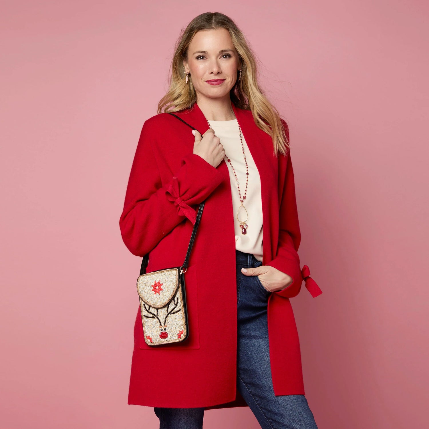 Woman wearing a red coat holding a decorative reindeer handbag against a pink background