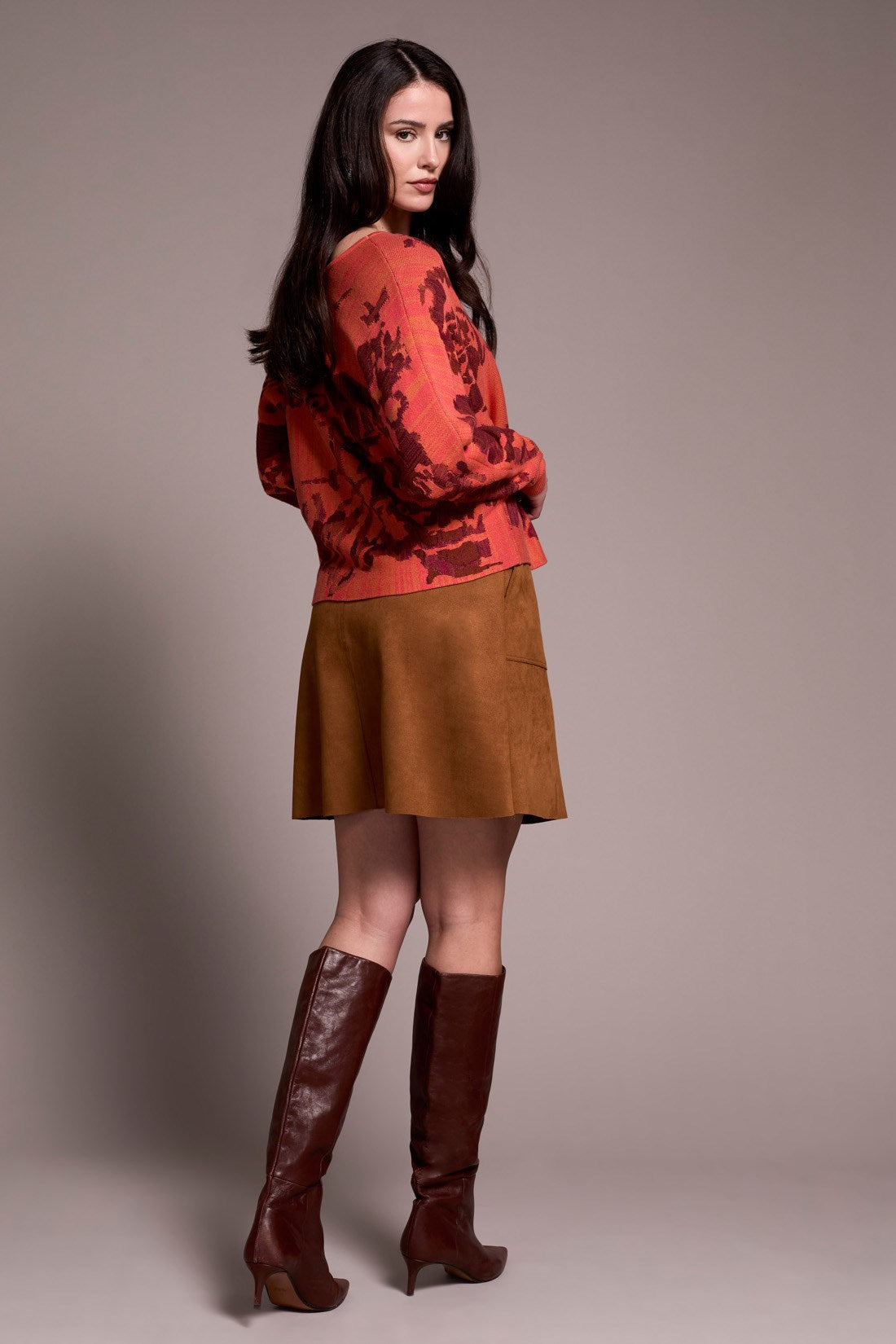 Woman wearing a burnt orange floral sweater, brown suede skirt, and brown knee-high boots against a plain background