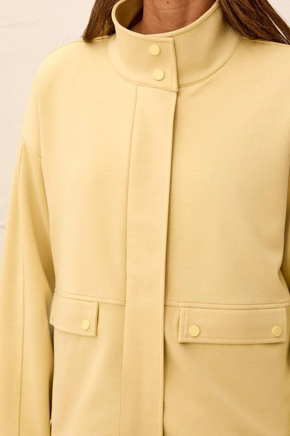 A close-up of a person wearing a pale yellow or butter-yellow jacket with a high stand collar, front button closure, and flap pockets with matching buttons tribal Soft-Touch Zip-Up Funnel Neck Utility Jacket in the color light pear