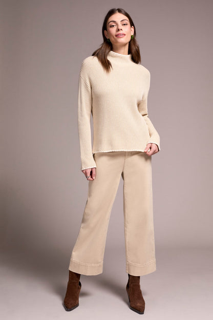 Woman wearing a beige sweater and pants with brown shoes on a plain background